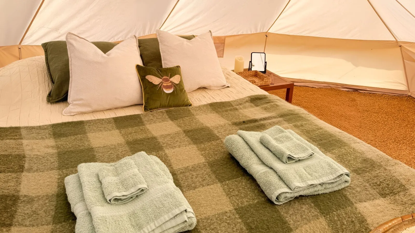 Inside a bell tent: a made-up double bed with a bee cushion, soft blankets and fresh towels on coir matting